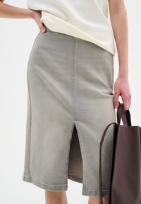 Fustă din denim cenușie, până la genunchi, cu o fanta pe spate, având cusături subtile și o textură netedă, purtată de o persoană care deține o genți maro.