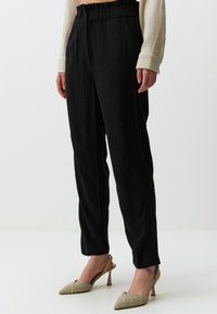 Schwarze, locker sitzende Hosen mit einem gerafften, elastischen Bund, kombiniert mit metallic-farbenen, spitzen hohen Schuhen. Glatte Textur mit einem dezenten Glanz.