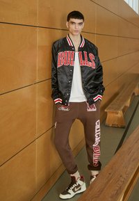 Svart satin varsity-jacka med röd "BULLS" text, ihop med bruna joggers med vita och rosa grafik. Högklackade sneakers fulländar looken.