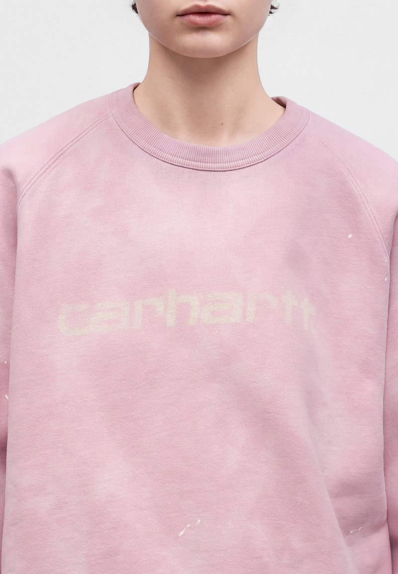 Person trägt ein hellrosa Sweatshirt mit Rundhalsausschnitt und schwachem "carhartt" Schriftzug über der Brust, zugeschnitten von der Nase bis zu den Schultern.