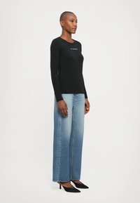 Vrouw staat in een zwarte langemouwen top met tekst, lichtblauwe wijde jeans en zwarte puntige hakken tegen een witte achtergrond.