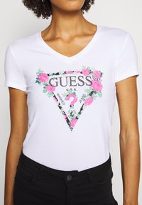Camiseta blanca de cuello en V con un logo triangular estampado de flores que incluye "GUESS" y acentos decorativos. Mangas cortas y diseño ajustado.