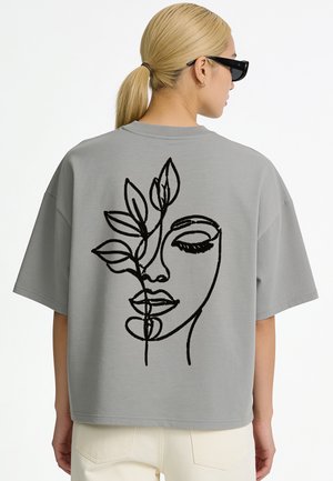 Blonde Person trägt graues oversized T-Shirt mit schwarzer abstrakter Linienkunst eines Gesichts und Blättern auf dem Rücken, kombiniert mit cremefarbenen Hosen.