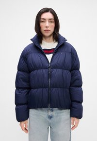 Granatowa kurtka puchowa z wysokim kołnierzem, pikowanym wzorem i zamek z przodu. Nosi ją na kremowym swetrze w czerwone i niebieskie paski.