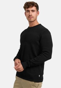 Maglione a maniche lunghe nera in maglia con righe orizzontali testurizzate, scollatura tonda e maniche con polsini. Il materiale sembra morbido e traspirante.