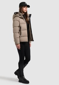 Chaqueta de plumas en taupe con costuras horizontales, bolsillos laterales con cremallera y capucha, usada sobre leggings negros y botas altas negras.