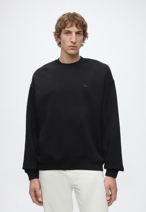 Junger Mann mit lockigem Haar, der einen schlichten schwarzen Pullover und weiße Hosen trägt, steht vor einem weißen Hintergrund.