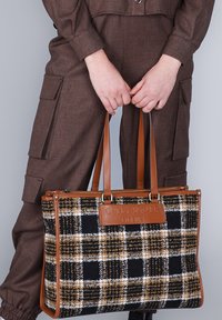 Ruce držící hnědou, černou a bílou kostkovanou tašku s koženými uchy a štítkem "CAFèNOIR FIRENZE", v kombinaci s hnědými cargo kalhotami.