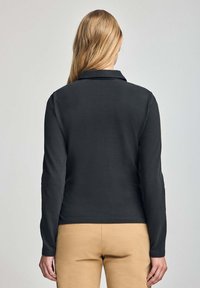 Camisa polo negra de manga larga con cuello, de tejido suave y diseño ajustado, combinada con pantalones marrón claro. Sin patrones visibles ni elementos metálicos.