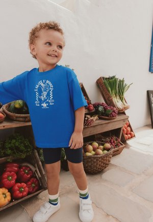 Lille barn med krøllet hår iført en blå t-shirt og shorts står smilende nær en markedstand med friske grøntsager og frugter.