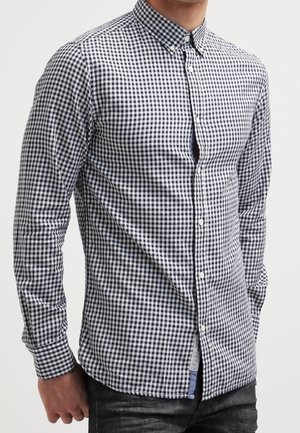 Mand iført en tætsiddende sort-hvid ternet skjorte med lange ærmer og mørke jeans mod en ensfarvet baggrund.