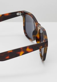 Sluneční brýle Vans v želvovinovém designu se tmavými čočkami, čtvercovým rámem, lesklým povrchem a logem na boku. Viditelné detaily kovových pantů.