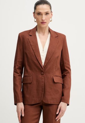 Kvinde med trukket hår tilbage iført guldringe øreringe, en rustrød blazer, matchende bukser og en hvid skjorte.