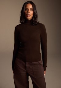 Jersey marrón de cuello vuelto y canalé con mangas largas, combinado con pantalones anchos de pierna en marrón oscuro con bolsillos laterales y acabado texturizado.