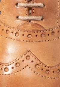 Tomaia in pelle marrone con dettagli brogue intricati, perforazioni, cuciture curve e una superficie liscia e lucida.
