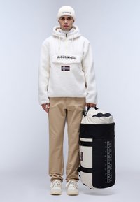Pullover bianco in pile con zip, logo e dettaglio della bandiera; pantaloni beige; sneakers bianche; borsa a secchiello nera e crema con logo.