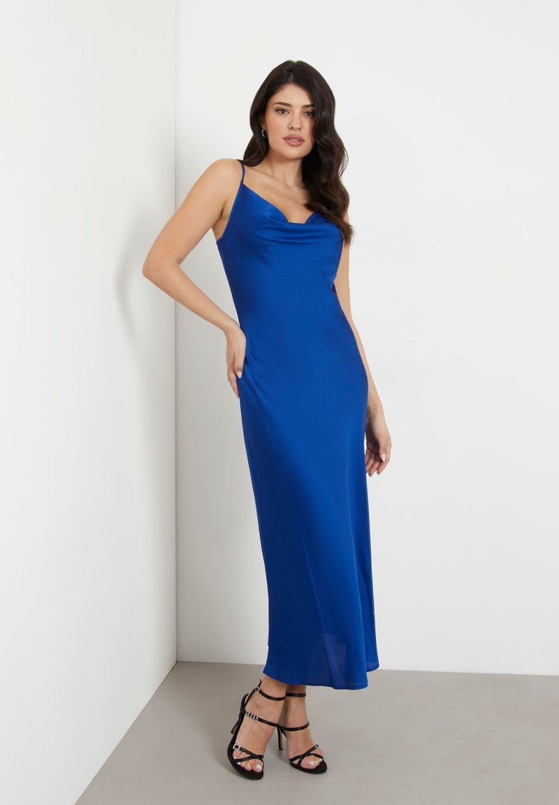 Woman standing in a blue sleeveless midi dress with cowl neckline and black strappy high-heeled sandals against a white wall.