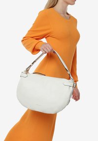 Bolso de hombro de cuero blanco con herrajes plateados, forma curvada y correa ajustable, sostenido por una persona que lleva un vestido naranja acanalado.