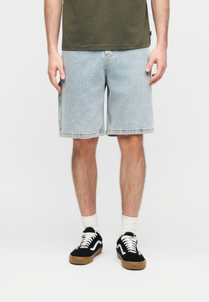 Άνδρας που φοράει ανοιχτό μπλε τζιν σορτς, ελιά πράσινο μπλουζάκι, λευκές κάλτσες και μαύρα αθλητικά παπούτσια με λευκά κορδόνια, στεκόμενος μπροστά σε απλό φόντο.