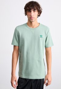 Žalias trumpas rankovėmis marškinėliai, pagaminti iš tekstūruotos medžiagos, su apvaliu kaklu ir mažu žaliu Adidas logotipu ant krūtinės. Atsitiktinė išvaizda.