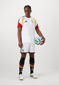 Λευκή φανέλα ποδοσφαίρου με κόκκινες και κίτρινες λεπτομέρειες, μαύρο σορτς και μαύρες κάλτσες με τη γερμανική σημαία. Ο παίκτης κρατά μια πολύχρωμη μπάλα ποδοσφαίρου.