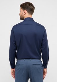 Camicia blu navy a maniche lunghe con collo strutturato e polsini con bottoni, caratterizzata da una texture liscia e una vestibilità sartoriale, abbinata a pantaloni azzurri.