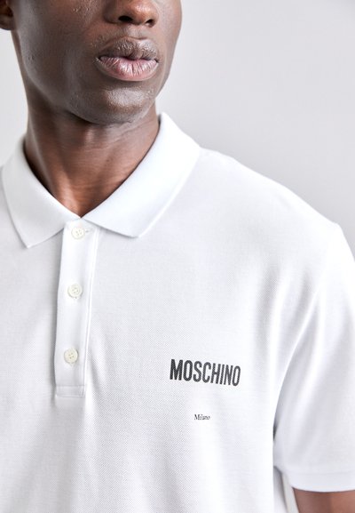 Valkoinen polo-paita, joka on valmistettu pintaneuloksesta, ja siinä on kolmen napin aukko sekä musta Moschino-logo, jonka alla on teksti "Milano".