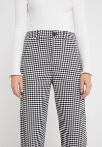 Donna che indossa pantaloni a scacchi bianchi e neri e una maglietta bianca a maniche lunghe con costine, mani rilassate ai lati su uno sfondo semplice.