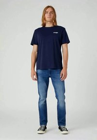 T-shirt in cotone blu navy con maniche corte, caratterizzata da un logo bianco sul petto, abbinata a jeans in denim blu chiaro e sneakers nere.
