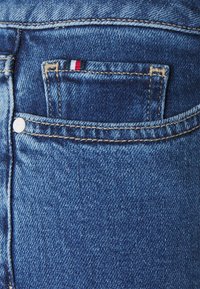 Tela de mezclilla azul oscuro con costuras claras en contraste. Presenta un pequeño bolsillo horizontal con un acento tricolor. Botón de moneda visible.