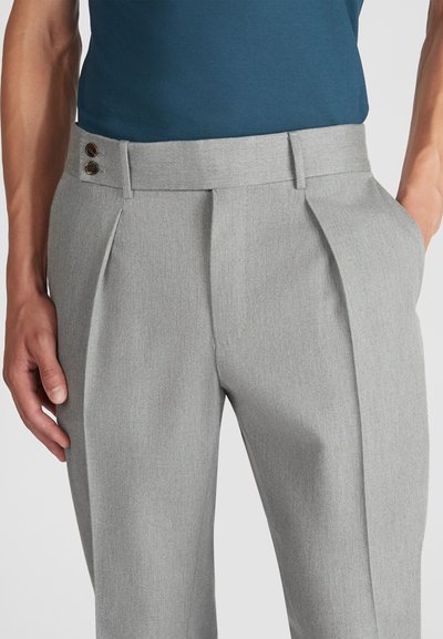 Man in een lichtgrijze geplooide broek met zijzakken en een dubbele knoopriem, gecombineerd met een donkerblauw shirt.