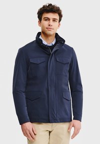 Granatowa kurtka wierzchnia z wysokim kołnierzem, dwiema kieszeniami na piersi i dwiema dolnymi kieszeniami. Wykonana z gładkiego materiału w dopasowanym kroju.