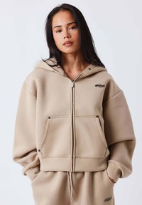 Sudadera beige con cierre de cremallera, ajuste holgado, capucha y bolsillos delanteros. Presenta un logotipo sutil y una textura de tela duradera.