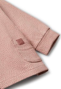 Softer rosa Stoff mit kleinen weißen Punkten, zeigt einen gefalteten Ärmel und eine kleine aufgenähte Tasche mit gesticktem Detail.