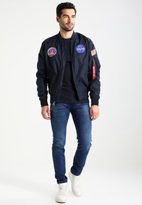 Marine bomberjakke med NASA-patches, lynlås foran og ribbede manchetter. Sammen med blå jeans og hvide sneakers. Glat stofstruktur.