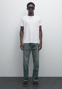 Mand iført sorte solbriller, en simpel hvid t-shirt, falmede straight-leg jeans og mørke sko, der står op ad en lys grå væg.
