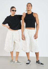 Two individuals stand side by side. The woman wears a black cropped shirt and white cargo shorts. The man has a black tank top and white cargo shorts. Both wear sneakers.