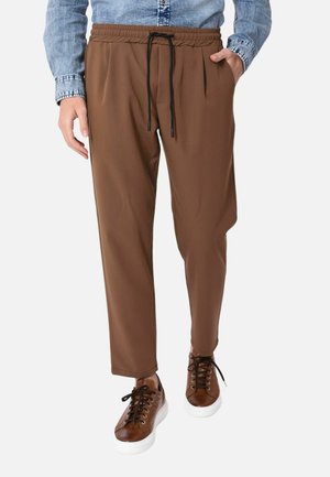 Uomo che indossa pantaloni marroni con coulisse e tasche laterali e sneakers in pelle marrone con suole bianche, abbinati a una camicia di jeans blu.