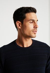 Maglione a maglia blu navy con texture a coste, scollatura rotonda e design aderente, che mette in risalto un tessuto liscio con lievi variazioni.