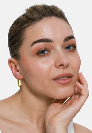 Boucles d'oreilles en or en forme de tube, portées par un modèle avec un maquillage neutre et un teint clair.