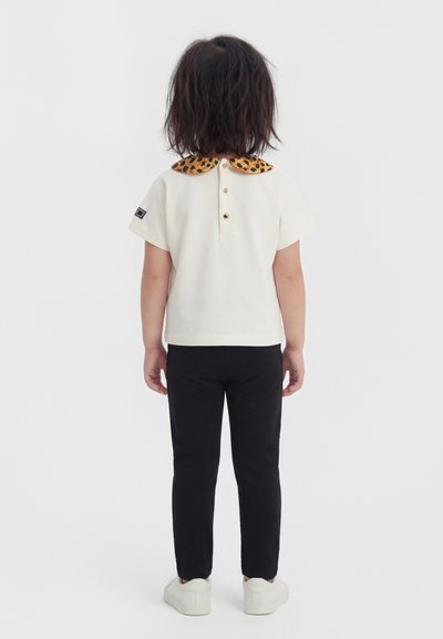 Enfant aux cheveux mi-longs debout de dos, portant un t-shirt crème à manches courtes avec un col à motif léopard, un pantalon noir et des chaussures blanches.