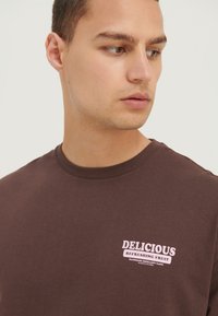 T-shirt en coton marron à manches courtes, col rond, avec un graphique en texte rose qui indique "FRUIT RAFRAÎCHISSANT DÉLICIEUX" sur la poitrine.