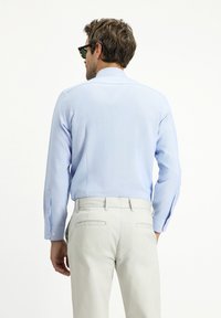 Camicetta azzurra a maniche lunghe con una texture discreta, polsini con bottoni e colletto, abbinata a pantaloni grigi chiari.