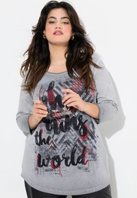 T-shirt gris à manches longues avec un motif graphique comprenant du texte et des motifs abstraits en noir, rouge et nuances de gris. Tissu doux, coupe décontractée.
