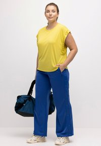 Top atletico giallo con maniche arrotolate e logo laterale, abbinato a pantaloni blu con bottoni a scatto e tasche con zip. Include borsa da palestra blu.
