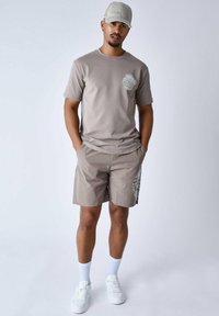 Camiseta de algodón marrón claro con estampado de logo, combinada con pantalones cortos a juego con un gráfico. Zapatillas blancas y calcetines completan el conjunto.