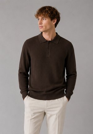 Homme aux cheveux bouclés portant un polo à manches longues marron foncé et un pantalon beige clair, mains dans les poches, regardant sur le côté.