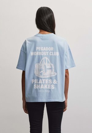 Person mit langen dunklen Haaren trägt ein hellblaues T-Shirt mit weißem Text und einer Illustration einer Sporttasche auf dem Rücken.