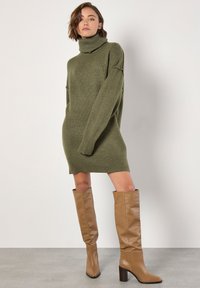 Vestido suéter de color verde oliva con cuello alto y puños y dobladillo acanalados. Combinado con botas de cuero marrón claro hasta la rodilla y tacón bloque.