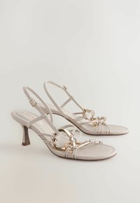 Helle beige High-Heelsandalen mit Kreuzband-Design, goldfarbigen Akzenten und einer glatten Textur sowie einer spitzen Zehenform.